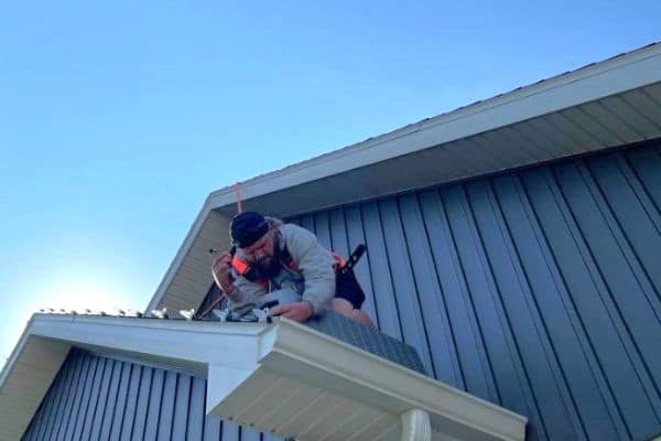Man working on a metal roof, secured with a safety harness, against a clear blue sky. He's installing new roofing or performing home repairs.