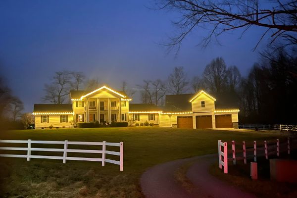 Large house adorned with warm white Christmas lights outlining the roofline at dusk. A white fence borders the lawn, leading to a driveway. Trees are silhouetted against the blue sky.