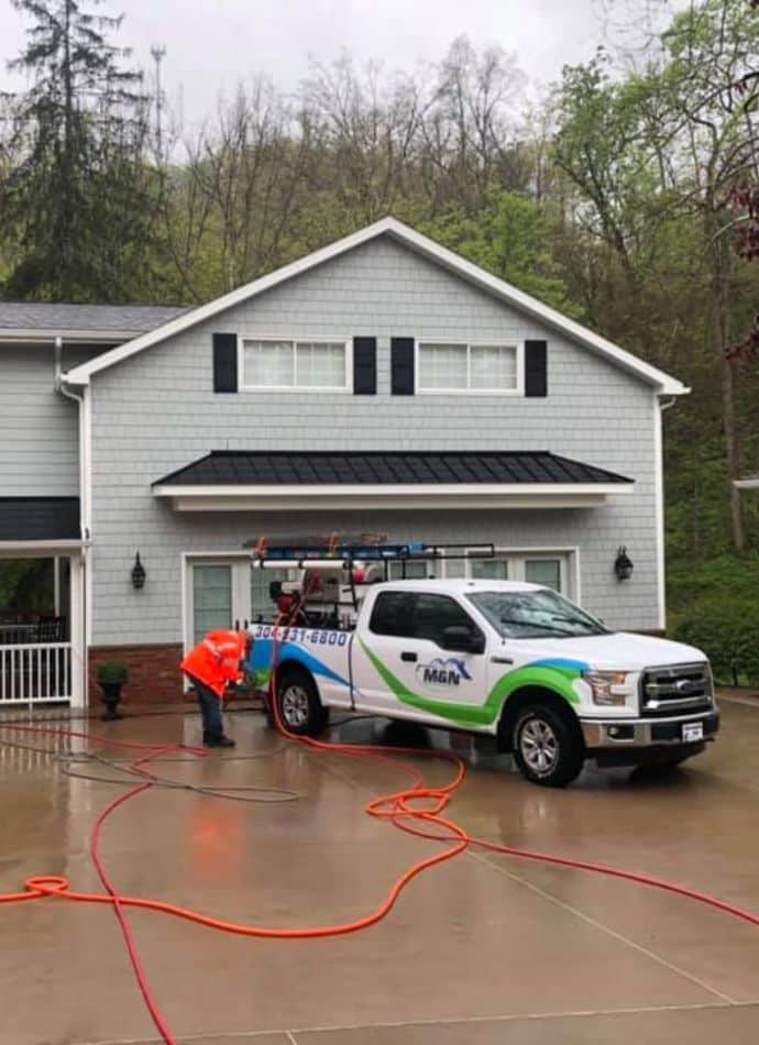 Power washing in progress: A worker in an orange vest power washes a driveway in front of a gray house with black shutters. A white truck with the logo "M&N" and the phone number 304-331-6800 is parked nearby.