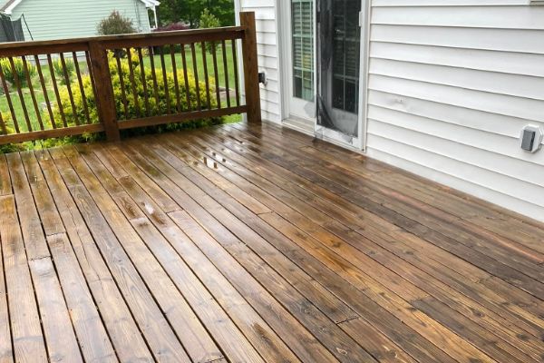 A freshly stained wooden deck gleams after the rain, showcasing rich brown tones. The deck is bordered by a white house and a green lawn, creating a cozy outdoor home space.