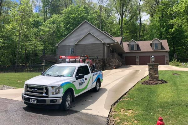 M&N Home Services truck parked on a driveway leading to a large house nestled in a green, wooded area. The truck has the company logo on the side.