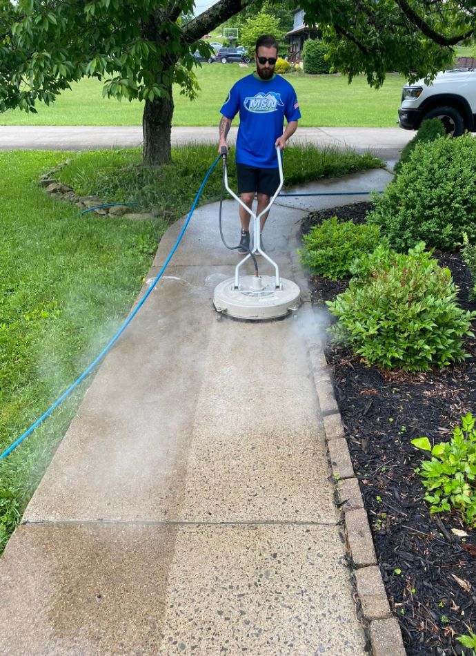 Man power washing a concrete sidewalk with a surface cleaner. He's wearing a blue shirt with "MSN" logo. Lush green lawn and landscaping border the walkway. Home maintenance.