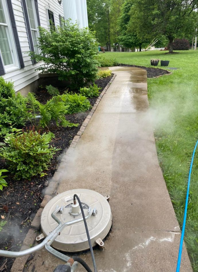 Power washing a concrete walkway next to a home. The cleaner reveals the bright concrete beneath dirt and grime, surrounded by lush green landscaping.