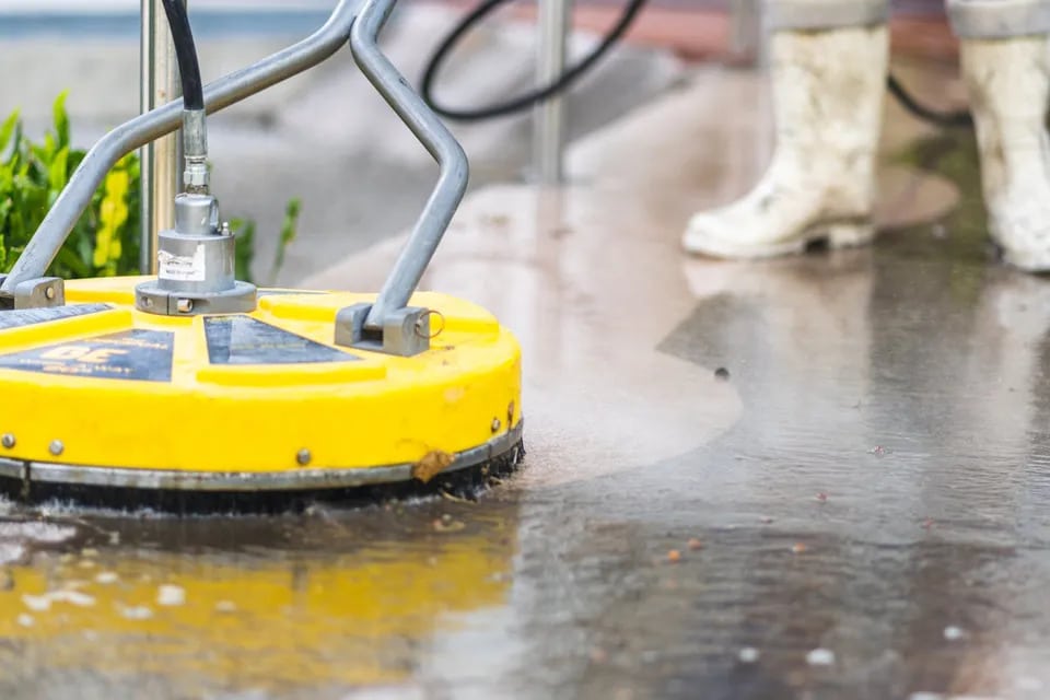 A yellow surface cleaner attachment on a pressure washer is used to clean a concrete surface, with a worker in white boots visible in the background. This image is relevant to pressure washing services.