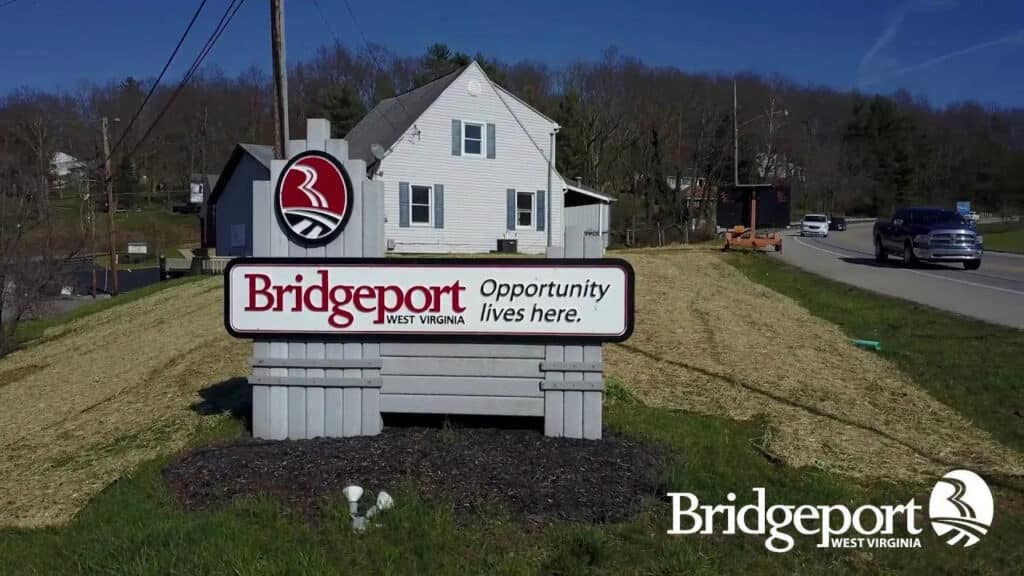 Bridgeport, West Virginia welcome sign featuring the city's logo and the motto "Opportunity lives here." A white house sits in the background beside a road with passing cars.