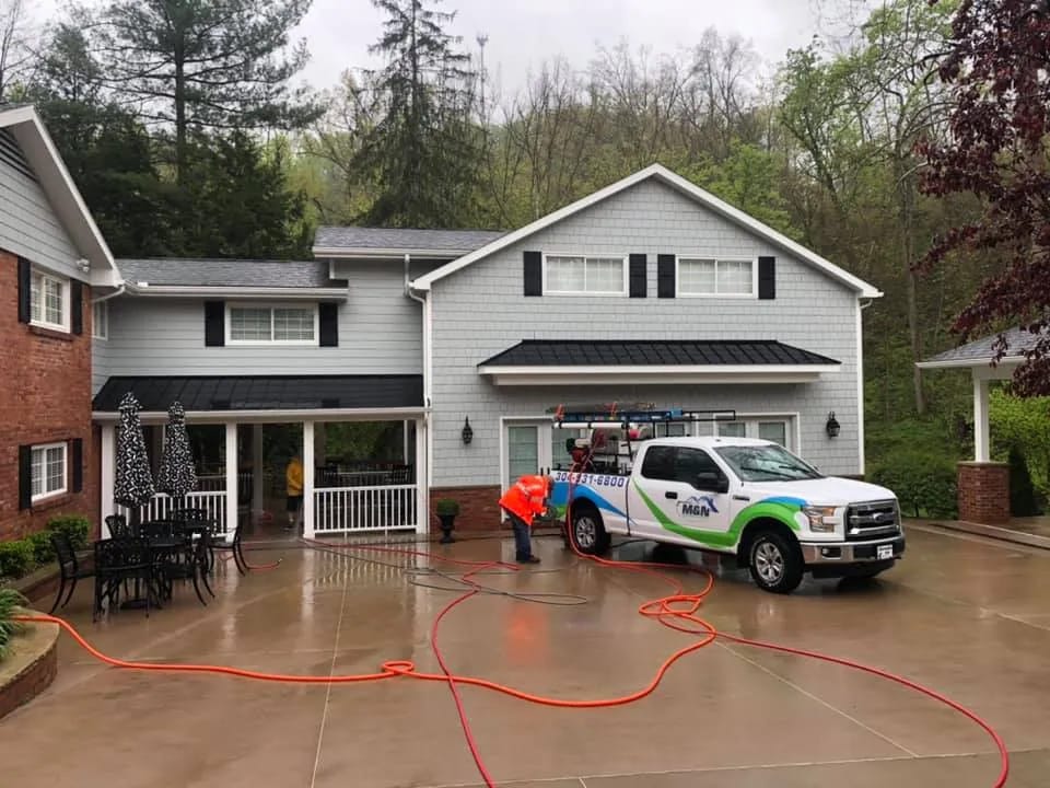 Soft washing in progress: A gray two-story house with black shutters is being cleaned by a worker with a pressure washer. A white M&N truck is parked in the driveway. Text on truck: 304-557-6500.