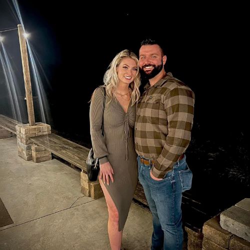 A smiling couple stands together outdoors at night. The woman wears a long-sleeved olive green dress, while the man is in a plaid shirt and jeans. They appear to be at home, posing for the photo.