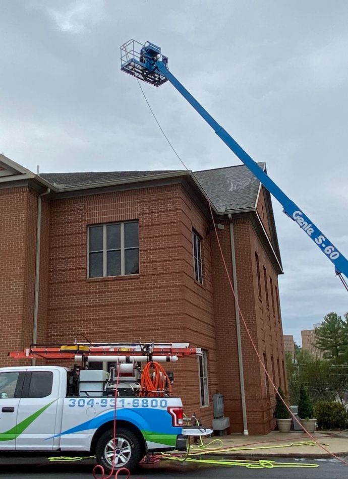 A Genie S-60 boom lift is extended to a brick building's roof for roof cleaning. A white truck with the phone number 304-931-6800 is parked below, likely related to the cleaning service.