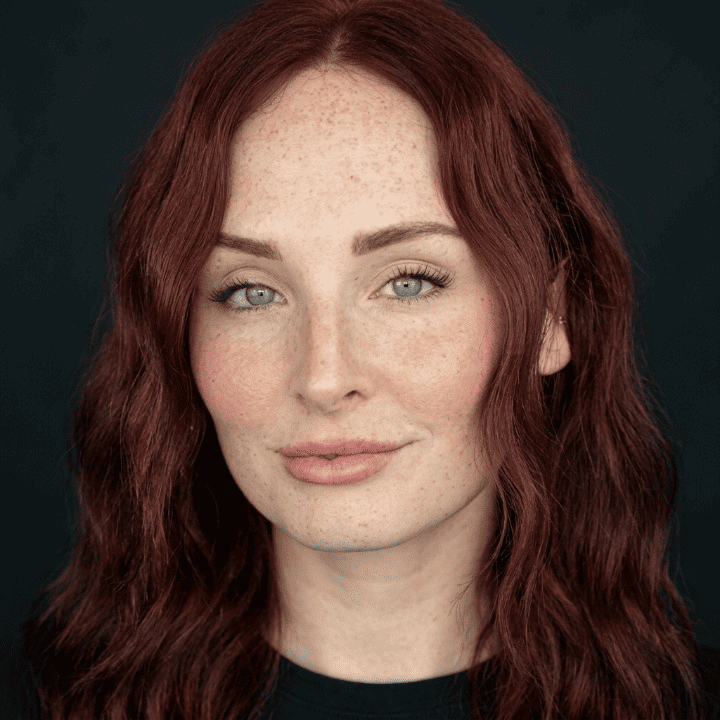 Close-up portrait of a woman with long wavy red hair, freckles, light blue eyes, neutral expression against a dark background.