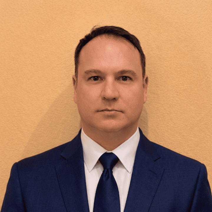 Head-and-shoulders portrait of a man in a blue suit, white shirt and navy tie against a beige textured wall, neutral expression.