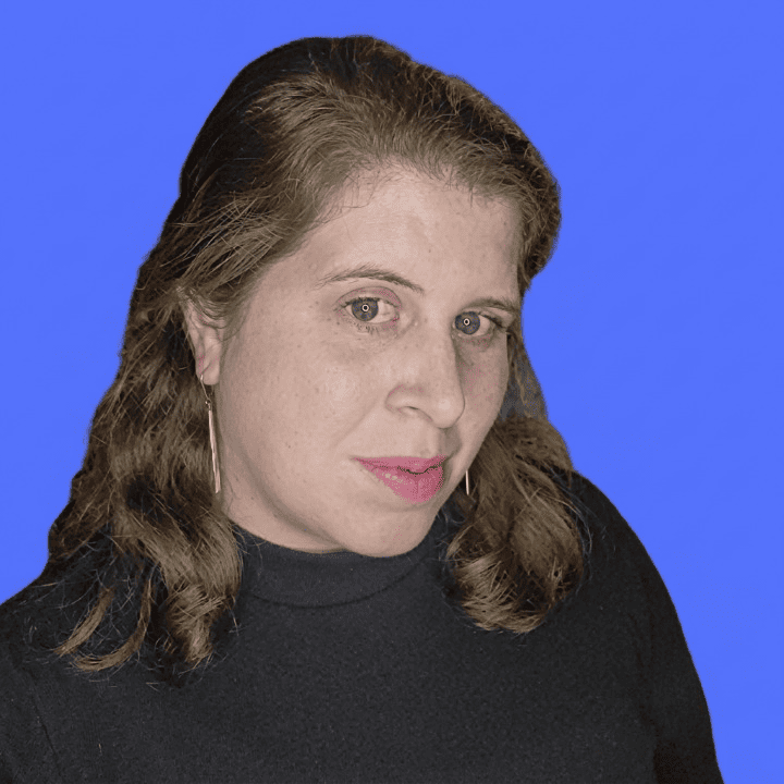Close-up of a person with shoulder-length brown wavy hair, pink lipstick, long thin earrings, wearing a black turtleneck on bright blue background.